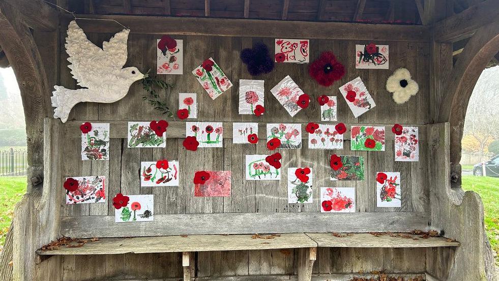 A bench with wooden shelter and back with a large dove of peace in one corner and childrens pictures of poppies, knitted ones scattered in between