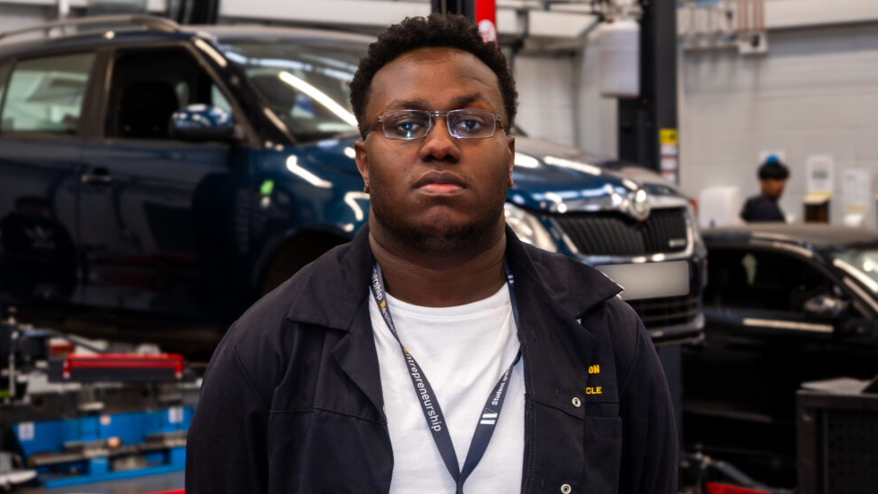 Antonio is wearing a white T'shirt and blue overalls and is stood inside a college work bay. There is a car raised up behind him with another close by. He has glasses on and tightly cropped hair.
