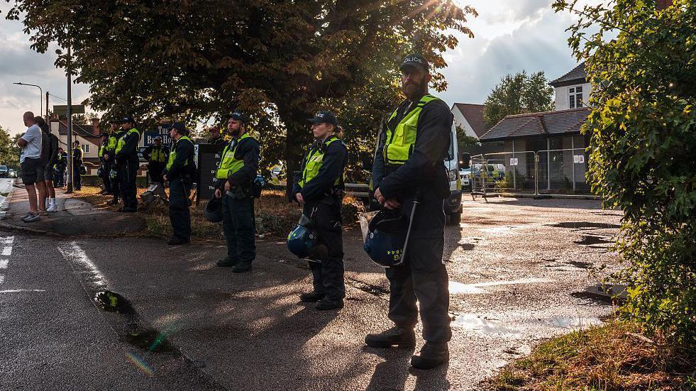 Police officers in black uniforms underneath green high-vis jacket stand in a line outside The Bell Hotel.