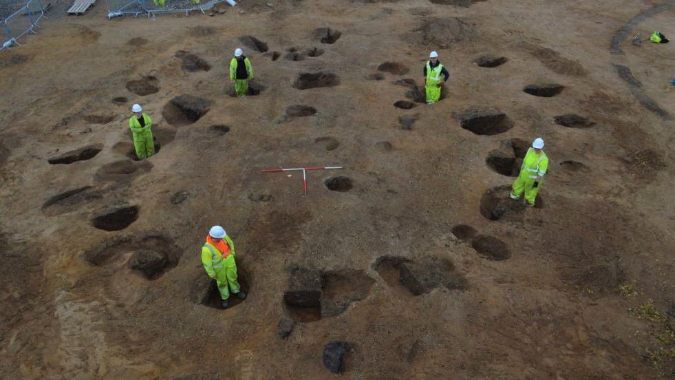 Archaeologists standing in post holes for the timbers of a roundhouse at an Iron and Bronze Age settlement in Inverness