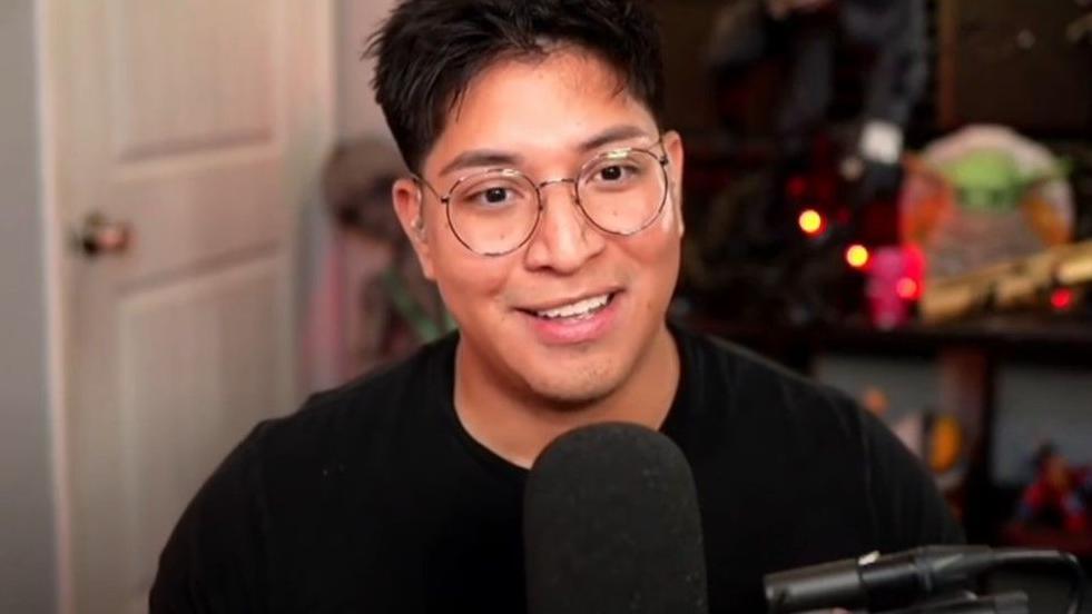 A streamer sits behind a microphone held in place on a boom arm. Behind him we can see shelves with various sci-fi and gaming-related merchandise lined up on them. He's smiling and wearing a plain black t-shirt