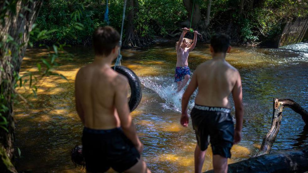 Three youths in shorts play on a rope swing over a river.