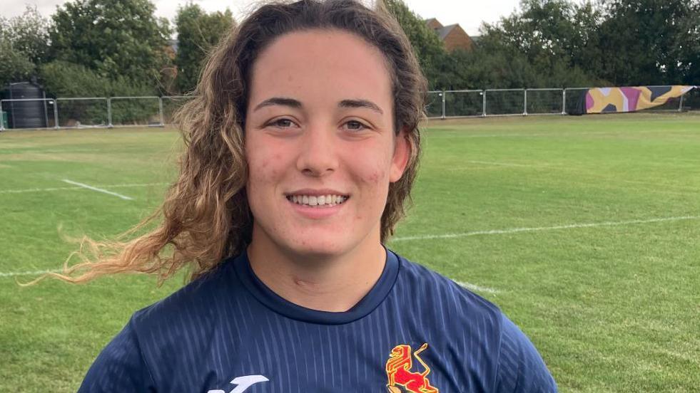 Alba Capell has long, wavy, brown hair and is wearing a navy sports top. She is smiling and standing on a rugby field.