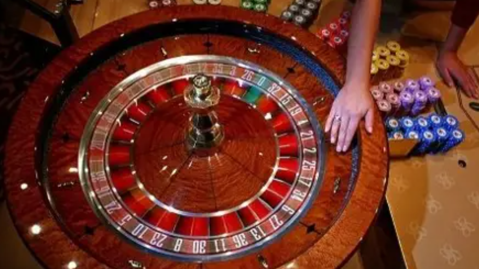 A roulette wheel, showing red and black numbers. There are chips in various different colours to the right of the table. A hand is spinning the wheel.