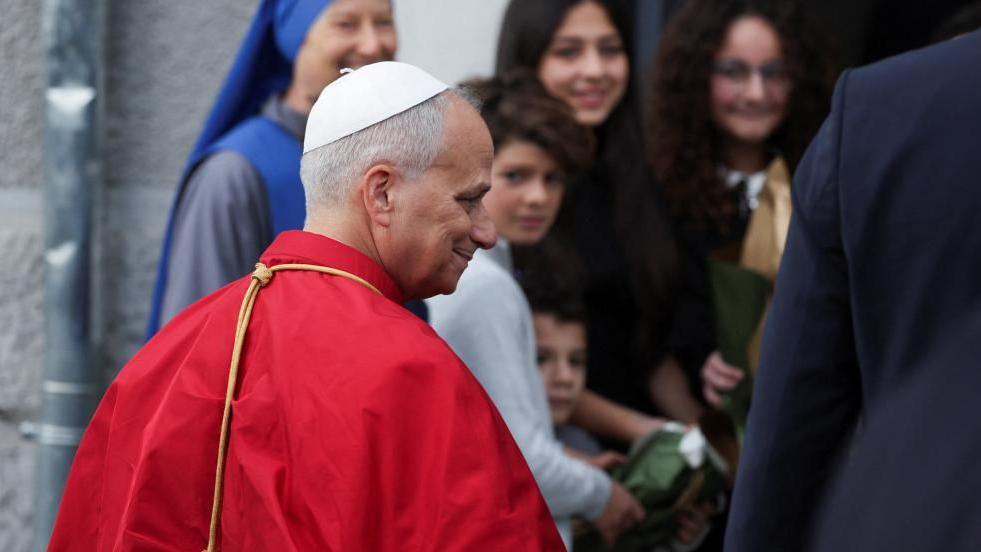 A man in a red mantle and white skullcap smiles outside a building where a small crowd of excited women and children stand.   
