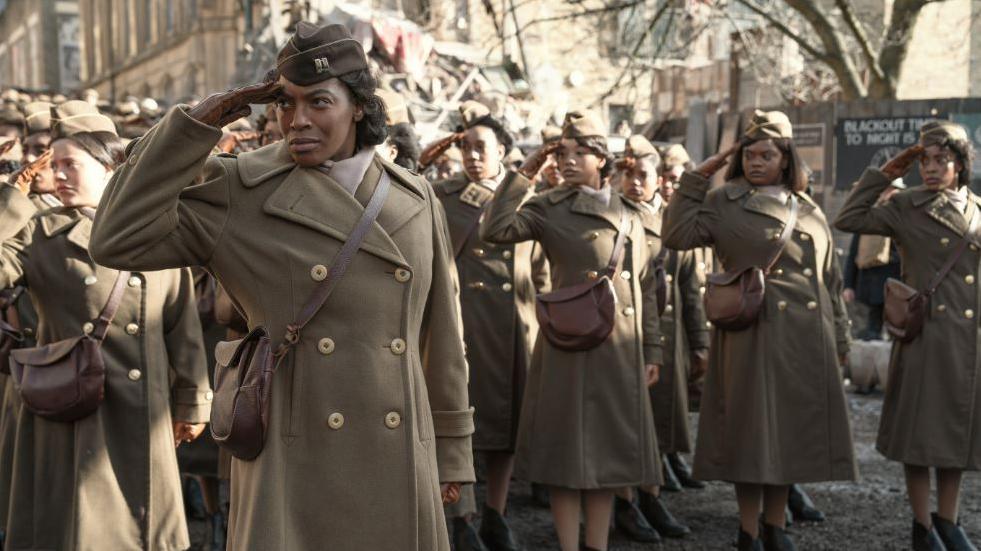 A still from the film. It shows rows of women in US World War Two uniform. they are all standing to attention, looking right and saluting  