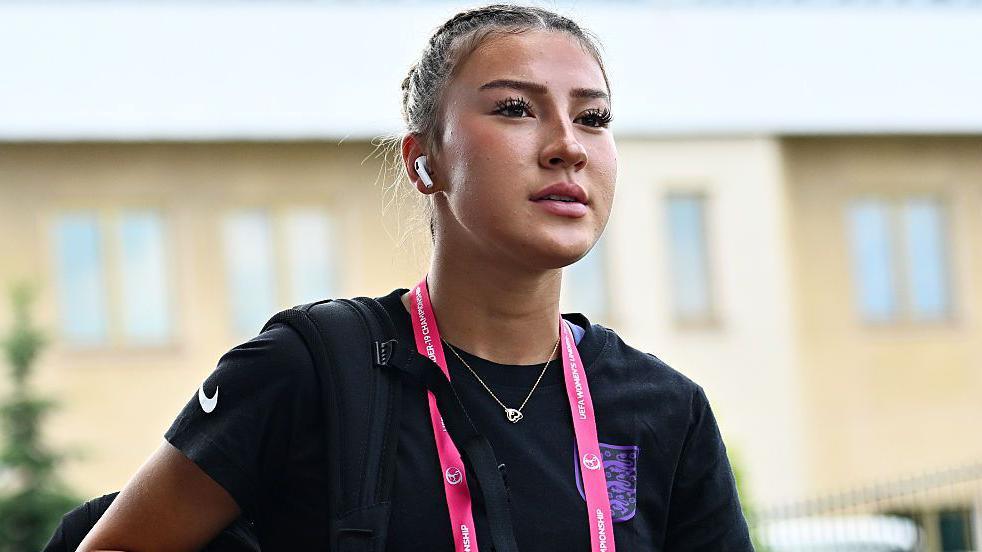 Erica wears a black t-shirt, a pink lanyard and has wireless headphones in her ears. 