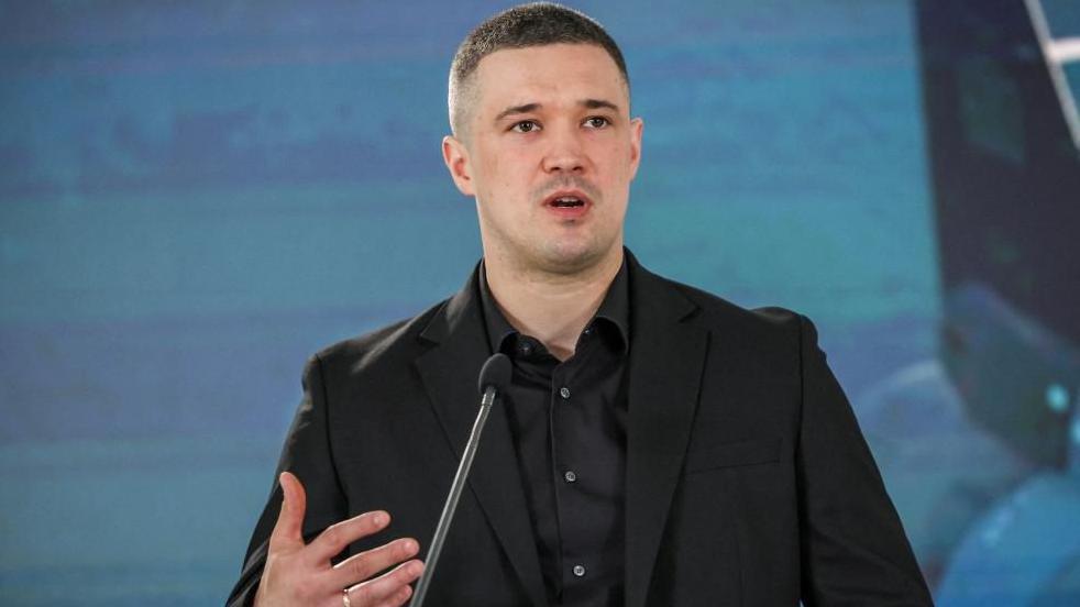 A man in a black jacket and black shirt talks in a microphone with a blue backdrop. His hair is close-cropped and he looks passionate.