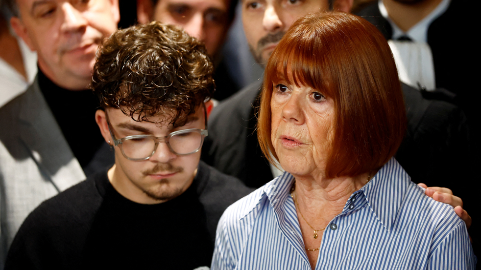 Gisèle Pelicot stands surrounded by male supporters, one of whom has his hand on her shoulder