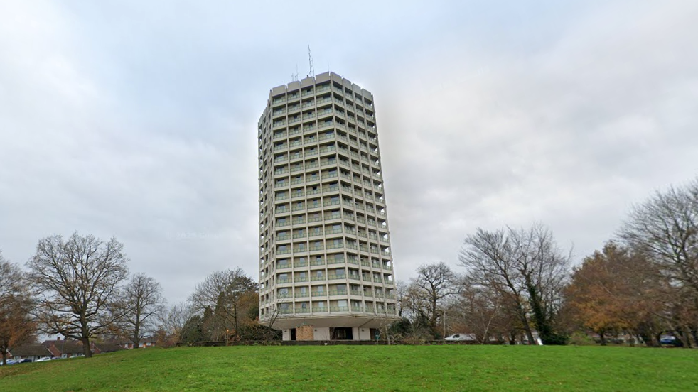 The 18-storey tower block Point Royal tower on Rectory Lane, Easthampstead. It is a large concrete hexagonal building with windows and balconies. It is surrounded by grass and trees. The picture is taken on a grey overcast day.
