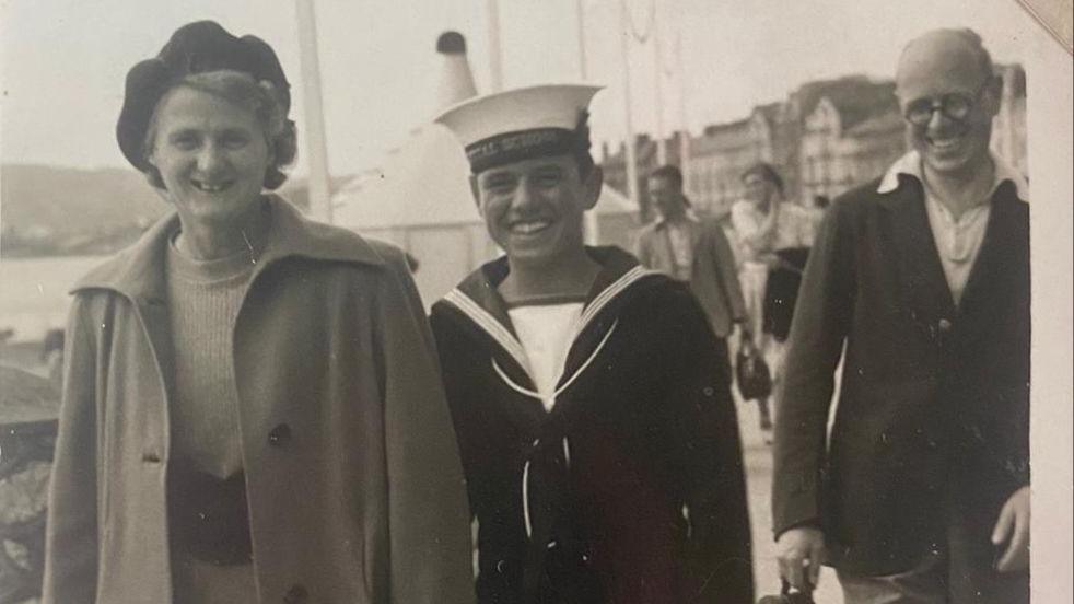 Black and white image of Derek Musselwhite in Navy uniform, smiling by a harbour. He is standing next to an older woman and a man, who are both also smiling. 