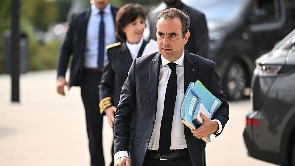Sébastien Lecornu walks with a folder and files through a car park while wearing a dark suit.