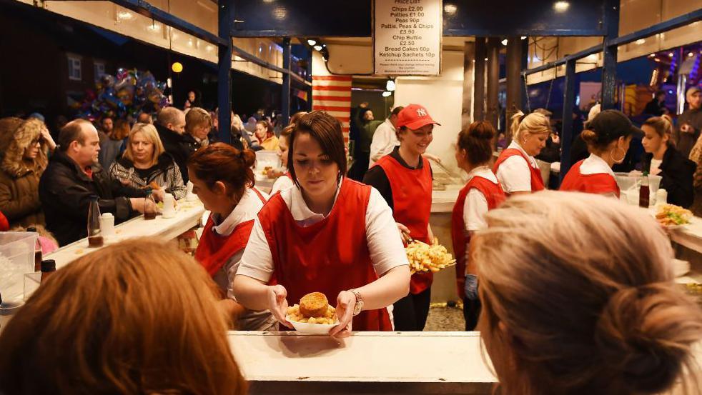A woman with a red apron over a white shirt is serving chips and a fried potato in batter to customers who are gathered around a blue and white stall.