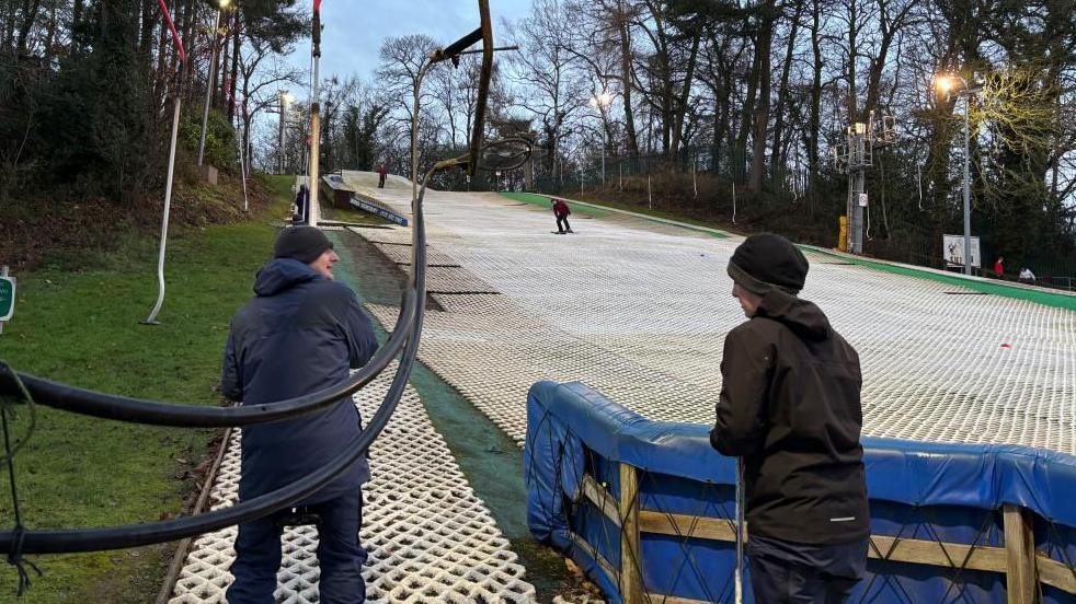 There is a ski slope made out of white bristles with green banking either side. There is also four skiers. The two closest to the camera are wearing dark coloured coats and black beanie hats. There is also a man in a red coat skiing half way down the slope. In the far distance, there is another man in a red coat getting ready to ski.