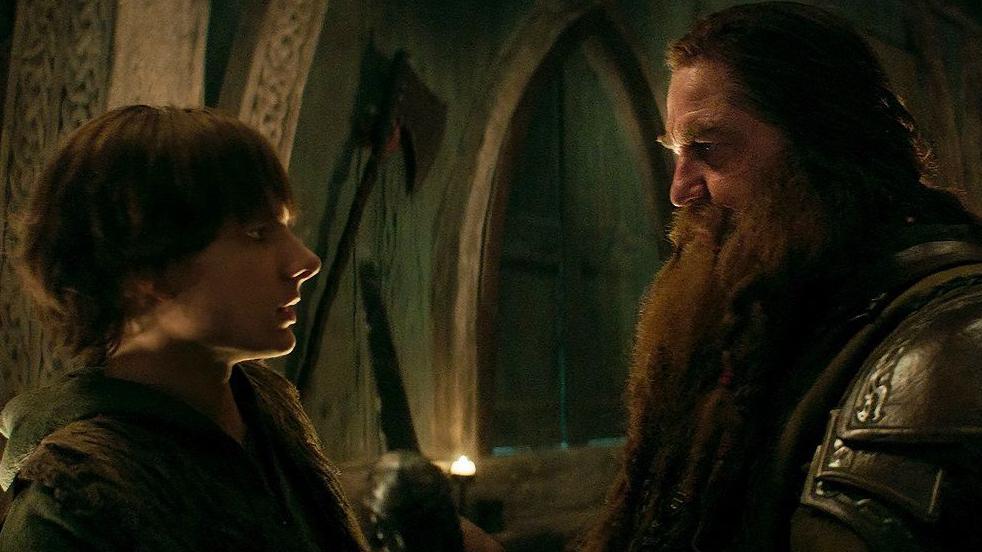 A young man and an an older man face each other - the young man has dark brown hair and is wearing thick wools. The older man has long dark hair, a long brown beard and is wearing armour. In the background are stone walls, with an axe hanging as decoration. It is a film set designed to look like it a Viking's home