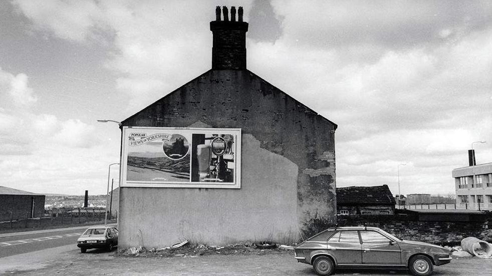 A black and white photo of a car parked in front of the gable end of a house with a beer advert on its side.