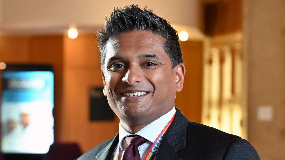 Dr Sandesh Gulhane smiles at the camera in the Scottish parliament. He has dark hair and wears a black suit with a white shirt and a burgandy tie. He also wear and NHS lanyard. 