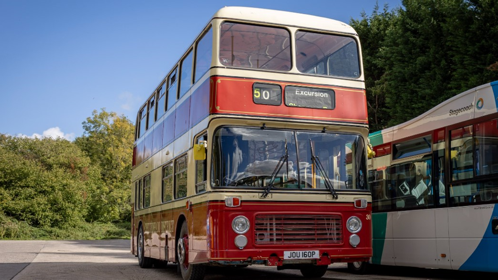 The red and beige striped bus is parked next to a modern-day bus with trees in the background and blue skies