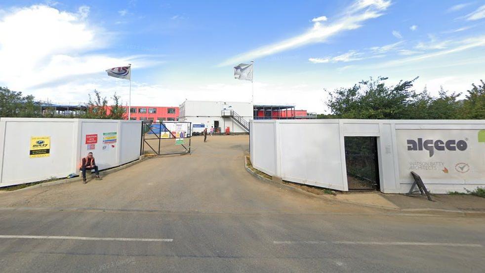 Construction site, showing white boards to screen the site. An entrance road with a black gate leads to a red school building.