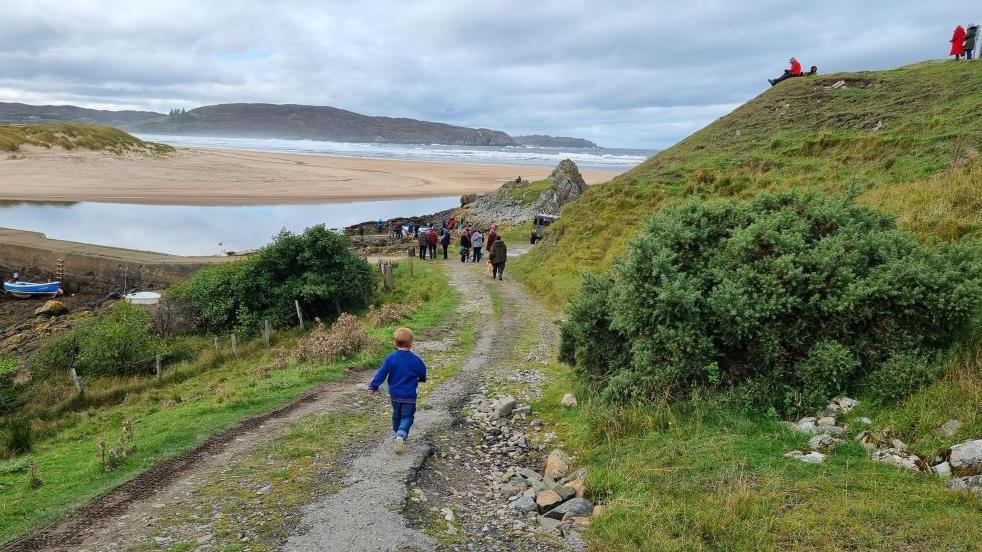 A boy runs down a track towards Bettyhill pier for the launch of the new boat. There is a crowd of people - and the beach - ahead of him.