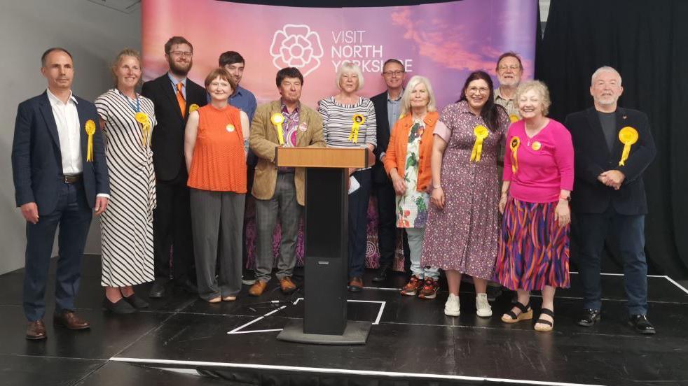 A collection of 13 people wearing yellow rosettes stand on a stage