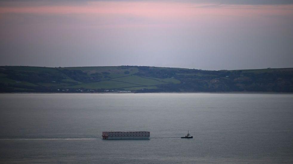 The Bibby Stockholm is seen from a distance, being pulled through the sea by a tugboat. In the background are country hills and a pink sunset.