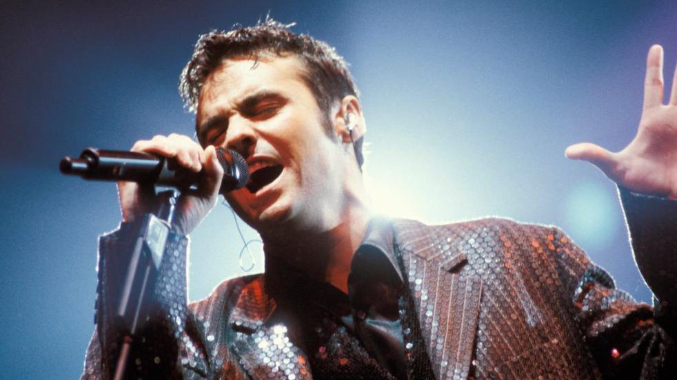 Marti Pellow from Wet Wet Wet singing into a microphone. He has short, dark hair and is wearing a shiny black shirt and a brown sequined jacket