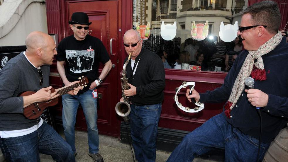 Image of four band members from the group Madness, wearing jeans and cardigans and performing on the street outside a pub. One band member is playing a ukulele, a second plays a saxophone and the lead singer Suggs has a microphone and tambourine. The other has no instrument but is standing, smiling.