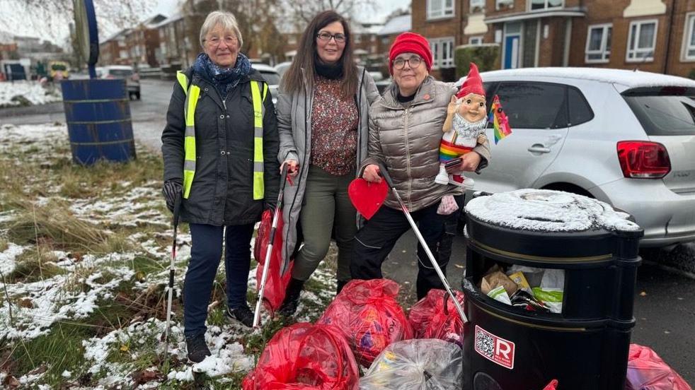 Three female volunteers are stood in a Blackburn street next to a bin surrounded with full bin bags. They are all dressed in winter clothing and are holding litter picker sticks. One woman is holding a garden gnome which is wearing rainbow shorts and holding a rainbow flag.