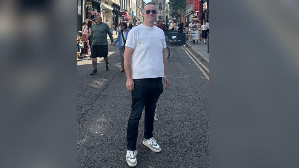 Martin Fletcher is standing in a street and is smiling while wearing sunglasses, a white top and dark trousers.