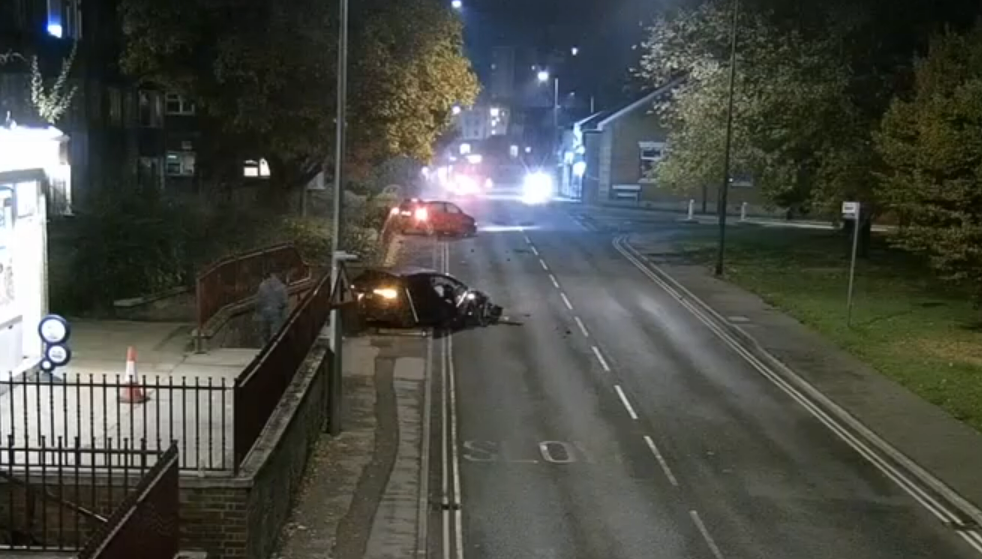 Crash scene in Ipswich. There is a shop on the left and a crashed black car can be seen on the pavement. Further away there is a red car which is also on the pavement in the image that was taken at night. 