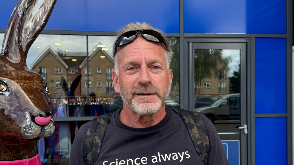 A head and shoulders picture of a man wearing a navy T-shirt and sunglasses on his head. He has a short beard and is looking directly into the camera. There is a large model of a hare just in shot on the left of the picture.