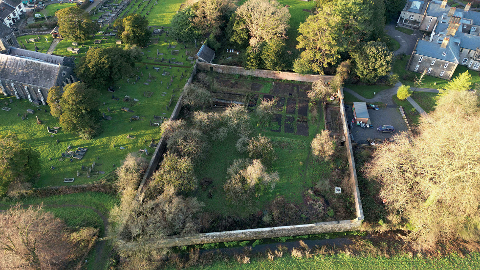 An aerial view of a square of grass surrounded by a grey brick wall, with less of it covered in vegetation than the previous photo. Three rows with narrow rectangles of dug earth can be see at the top of the square. 
