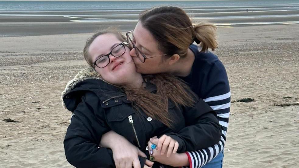 Emily McAllister, wearing a navy jumper, hugging and kissing her daughter Keira, who has down syndrome and is wearing a black coat, as they stand on a sandy beach. 