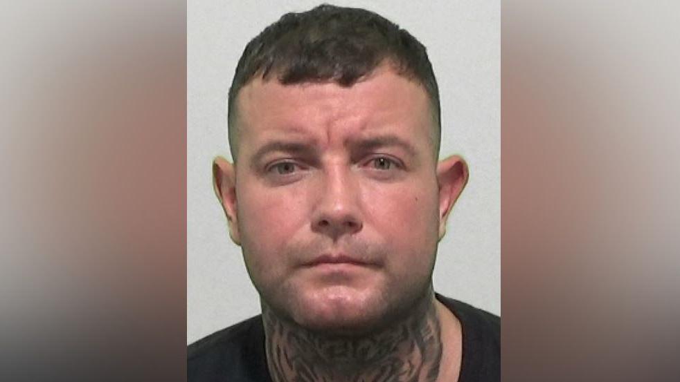 Mugshot of Hazard. He has a large tiger's face tattooed across the entirety of the front of his neck, is clean shaven with short dark hair.