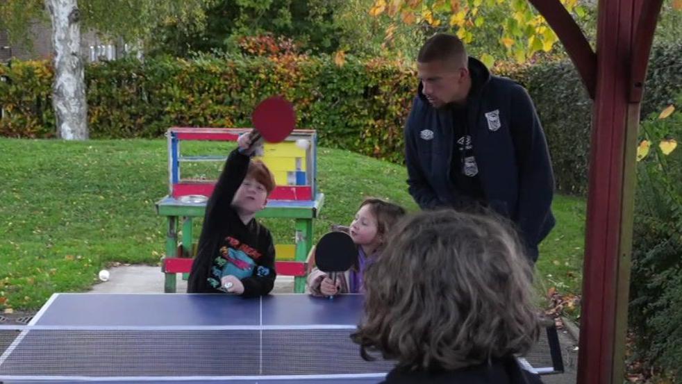 Footballer Harry Clarke plays table tennis with children outside. Two children stand beside him holding bats with another child standing opposite with their back to the camera.
