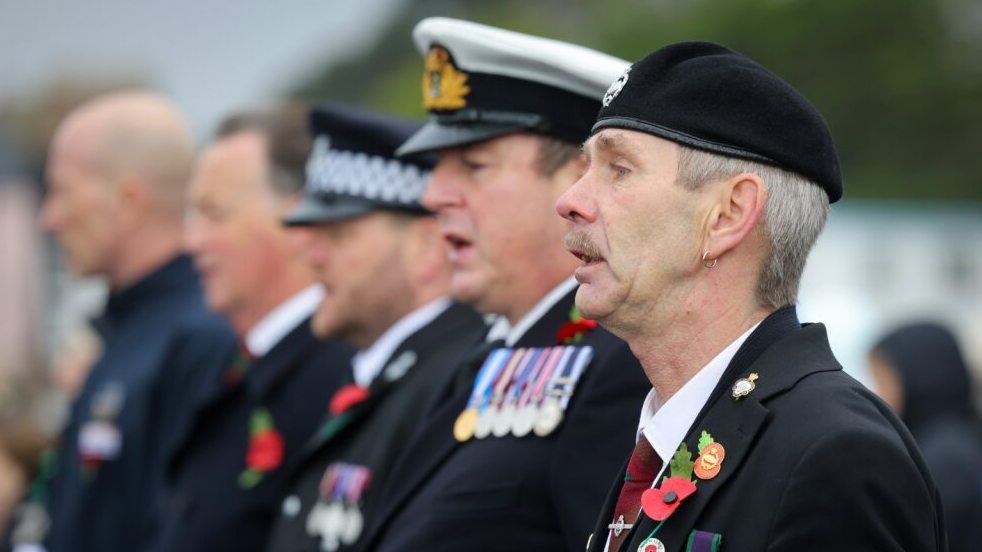 A line of men take part in the service.  The four men are dressed in black uniforms. They are wearing poppies on their clothing as well as badges and medals. Their mouths are slightly open which suggests they are talking or singing. 