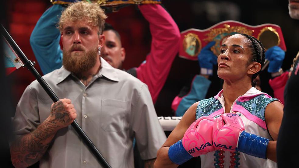 Jake Paul holding a flag pole and standing next to Amanda Serrano who is pushing her boxing gloves together