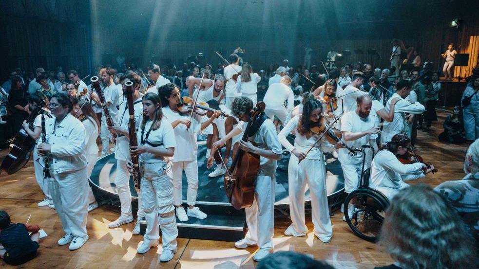 An orchestra playing on a circular stage, all dressed in white, some standing and some in wheelchairs