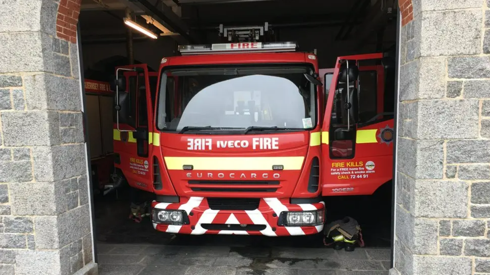 The front of a red fire engine with its doors open inside of a building. Either side of the fire truck is a light-coloured stone wall.