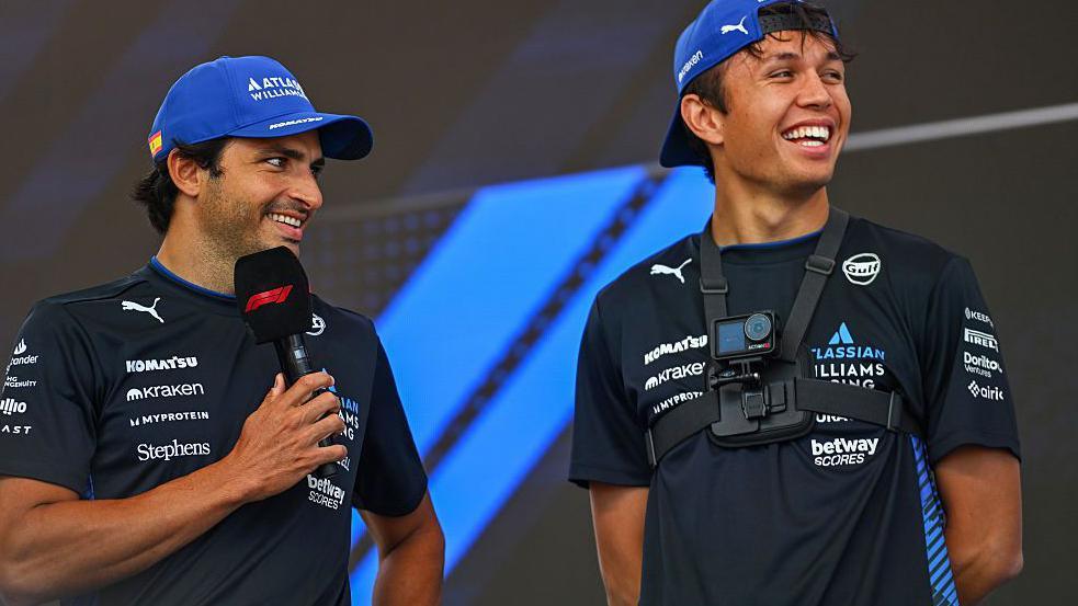 Carlos Sainz holds an F1-branded microphone with Alex Albon stood next to him. Both are wearing Williams t-shirts and caps
