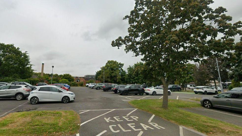 A view of a car park full of vehicles. There is a large tree in the foreground and a "Keep clear" sign is painted on the ground.