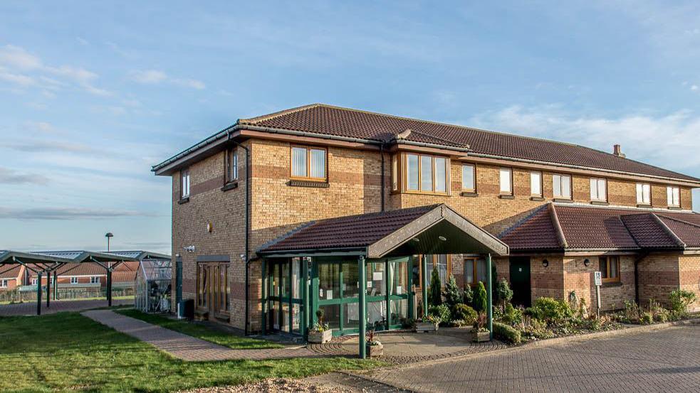 The outside of Lakelands Hospice, which is a large two-storey brick building with a green porch entrance.