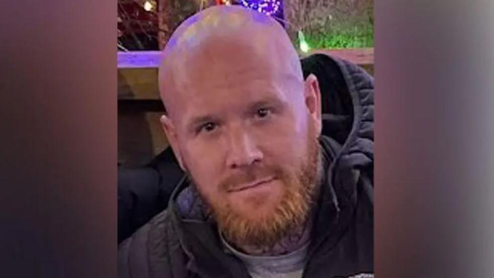 Lenny Scott, who has brown eyes, shaven head and ginger beard and is wearing a navy blue puffer coat and grey t-shirt.