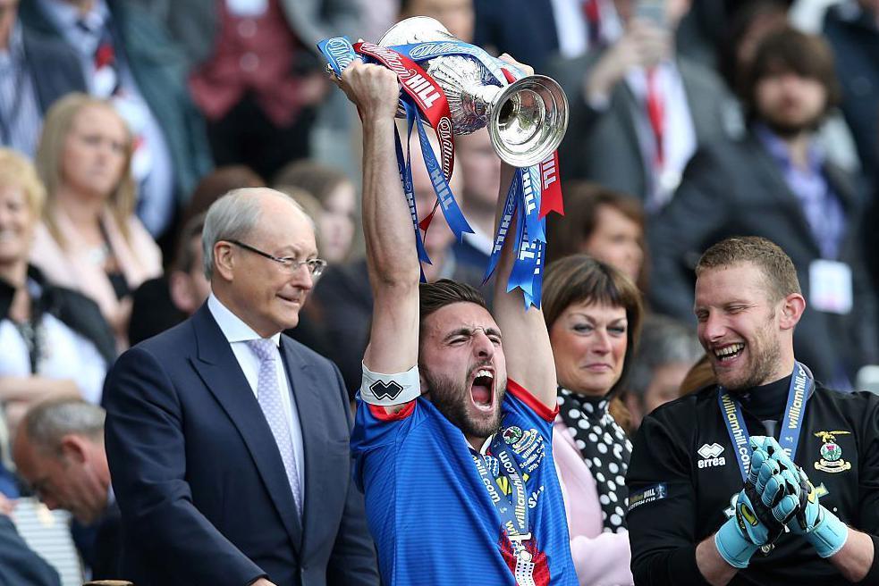 Graeme Shinnie holds aloft the Scottish Cup. Team-mate, goalkeeper Ryan Esson, looks on smiling. 