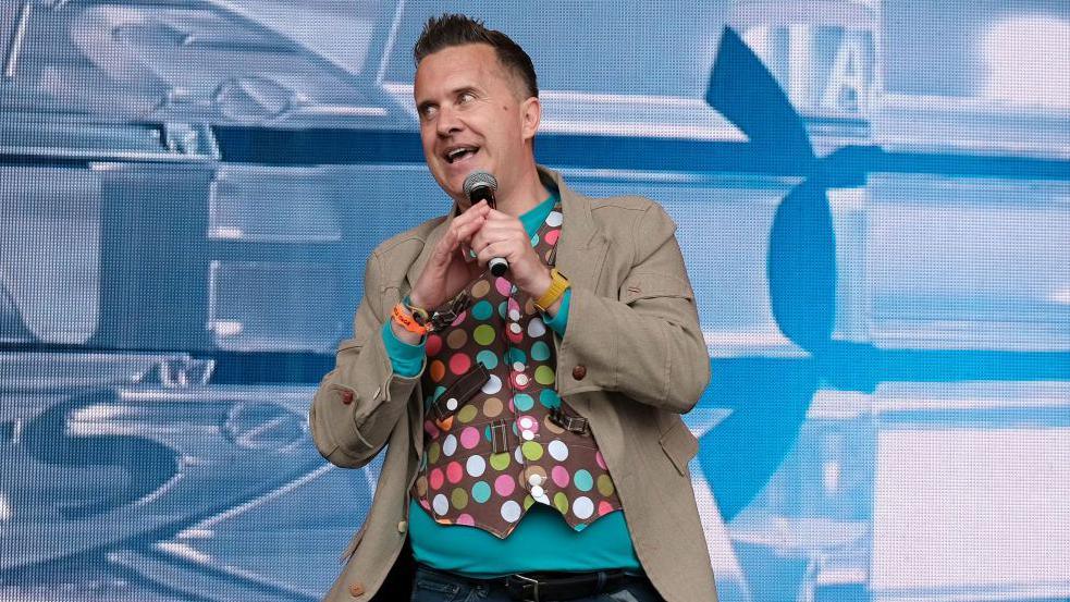 Mister Maker performing live on stage at Camp Bestival. He is wearing a light jacket with a multi-coloured polka dot waistcoat underneath.