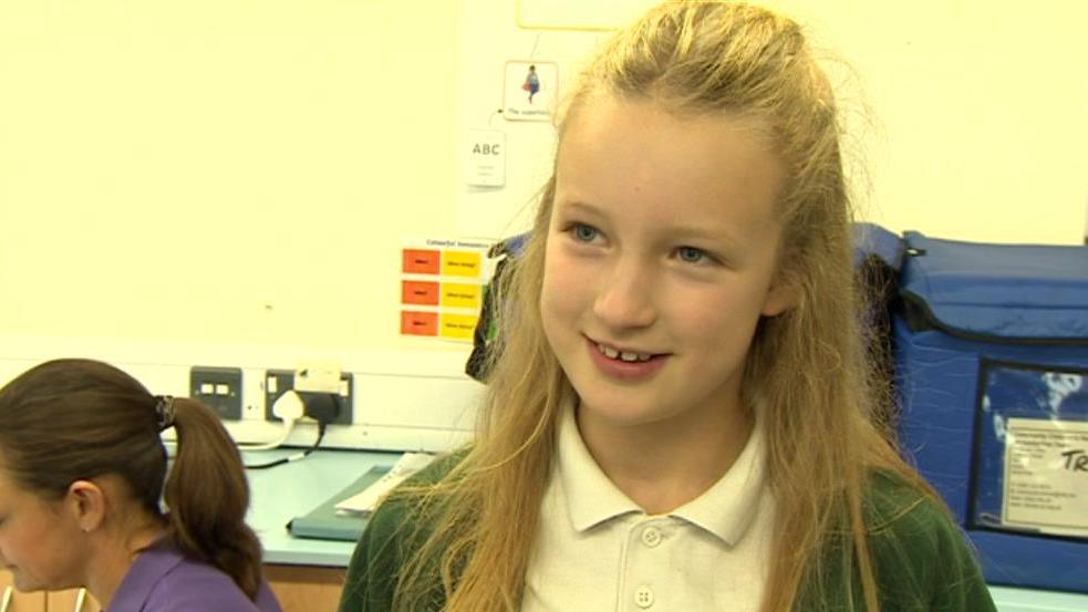 A blond girl of primary school age wearing a white polo shirt and green jumper looks at the camera.  A nurse sits at a desk behind her. 