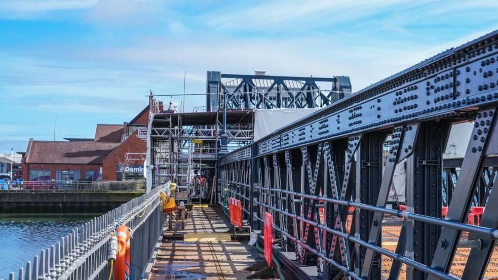 A bridge with a low railing on the left and a high railing on the right. There is scaffolding at the far end.