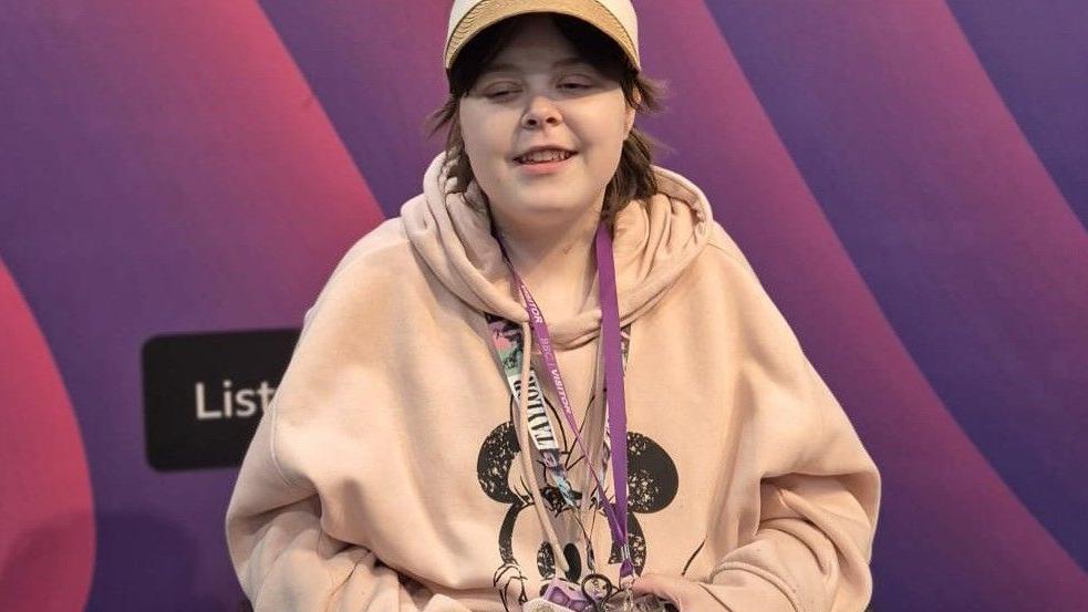 Lilly in a radio studio. She is wearing lanyards. She has a beige Micky Mouse hoody on and a beige hat.