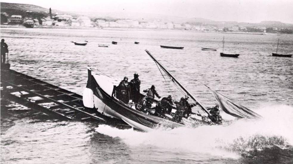 The Swanage lifeboat Zaida (1914-1918) crashes into the sea from a slipway throwing up a large spray of water. Zaida is an open boat with collapsed sails and at least seven crew on board. Photo is black and white.